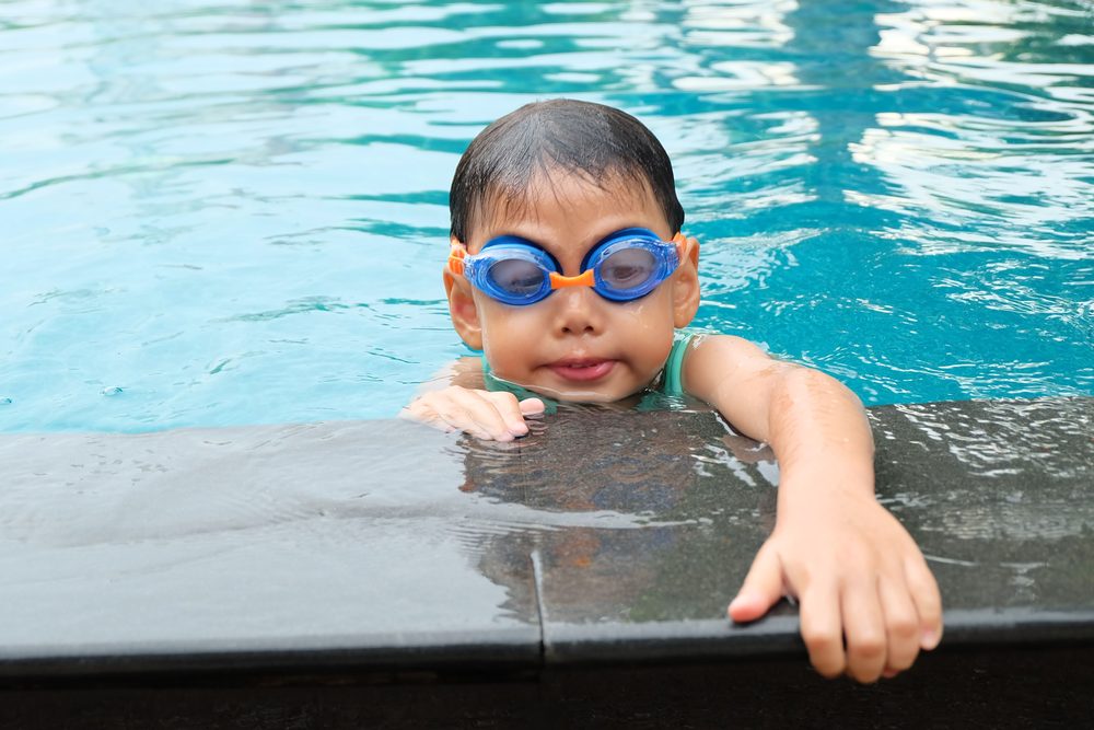 A young boy in goggles holding the edge of a pool, smiling, partway through a swim lesson.