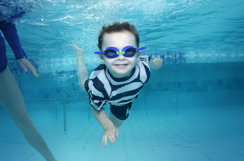 A young boy in goggles swimming underwater towards the camera during a lesson, an instructor beside him.